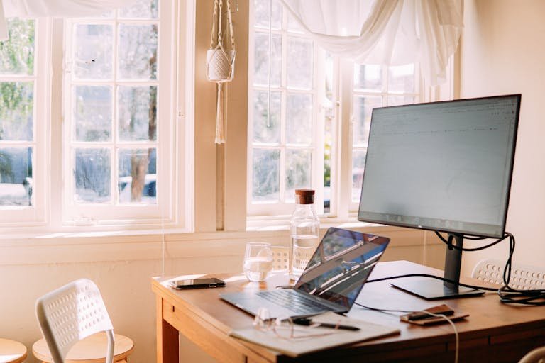 A sunlit home office space featuring a laptop on a wooden desk with natural light streaming through large windows.